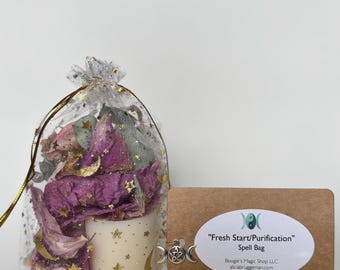 Op de afbeelding: Een spreukzak met een witte kaars versierd met gouden sterren en maan. De zak bevat gedroogde bloemen en een label met de tekst "Fresh Start/Purification" Spell Bag.