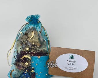 May include: A blue mesh bag filled with dried flowers and herbs, with gold stars and crescent moons. A turquoise candle with gold stars is next to the bag. A brown tag with the text "Healing Spell Bag" is attached.