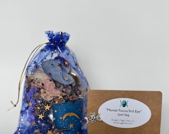 Op de afbeelding: Een blauwe mesh zak gevuld met gedroogde bloemen en een kleine blauwe kaars met gouden sterren en een maanontwerp. Een bruin label met de tekst "Mental Focus/3rd Eye" is bevestigd met een bedel.