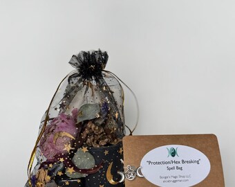 Op de afbeelding: Een zwarte mesh tas gevuld met gedroogde bloemen en kruiden, vastgebonden met een zwart lint. Een kleine kaart met de tekst "Protection/Hex Breaking Spell Bag" en een zilveren bedel met een maan en ster zijn ook aanwezig.