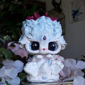 May include: A white and light blue fantasy creature figurine with large black eyes and a purple gem on its forehead. The figurine has a textured, scaled appearance and is surrounded by white and pink flowers.