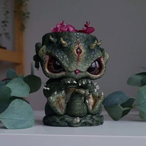 May include: A detailed, green dragon-like planter with large, expressive eyes and small horns. The planter is filled with pink flowers. The dragon has a textured, scaly appearance and is sitting down. A unique, fantasy-themed decorative item.