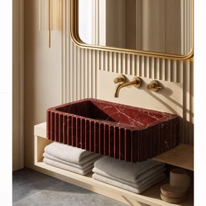 May include: A modern bathroom sink made of red marble with white veining. The rectangular sink has vertical grooves and is mounted above a shelf with folded white towels. A gold faucet and mirror frame complement the design.