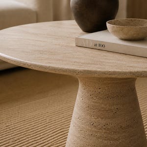 May include: A round, beige stone coffee table with a pedestal base. A dark brown vase, a small beige bowl, and a book with the text "at 100" are on the table. The table is on a beige rug.