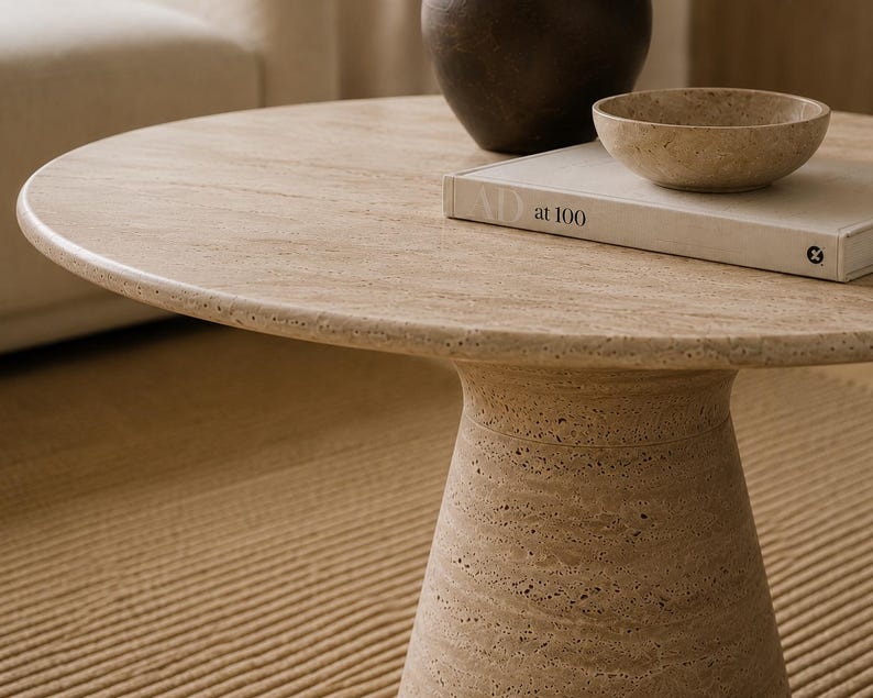 May include: A round, beige stone coffee table with a pedestal base. A dark brown vase, a small beige bowl, and a book with the text "at 100" are on the table. The table is on a beige rug.