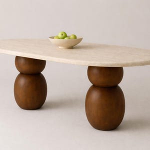Op de afbeelding: Ovale salontafel met een lichtbeige blad en twee bruine, gestapelde, afgeronde poten. Een kleine, beige kom gevuld met groene appels staat op de tafel. Modern design.