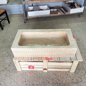 May include: A rectangular, light beige stone bathroom sink with three faucet holes. The front of the sink has a textured, wavy design. The sink is resting on a wooden crate with red warning labels.