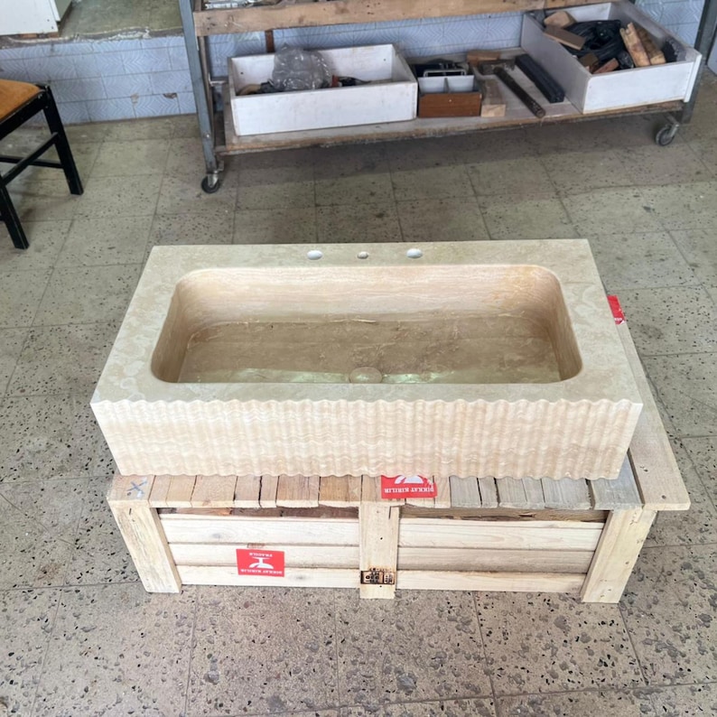 May include: A rectangular, light beige stone bathroom sink with three faucet holes. The front of the sink has a textured, wavy design. The sink is resting on a wooden crate with red warning labels.