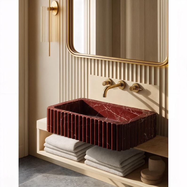 May include: A modern bathroom sink made of red marble with white veining. The rectangular sink has vertical grooves and is mounted above a shelf with folded white towels. A gold faucet and mirror frame complement the design.