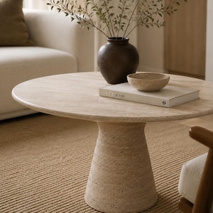 May include: A round, beige travertine coffee table with a pedestal base. A dark brown vase with greenery, a small bowl, and a book are on the table. The table is in a living room setting with a beige sofa and rug.