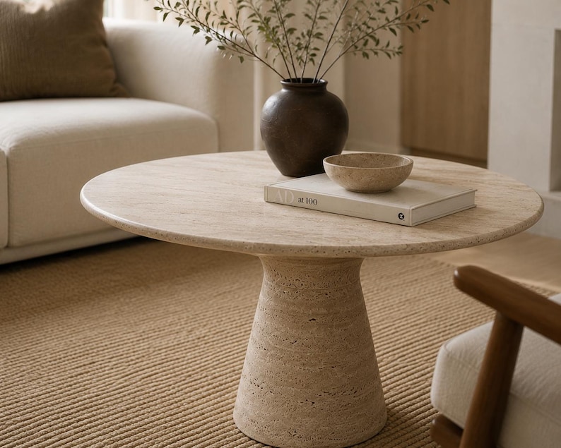 May include: A round, beige travertine coffee table with a pedestal base. A dark brown vase with greenery, a small bowl, and a book are on the table. The table is in a living room setting with a beige sofa and rug.