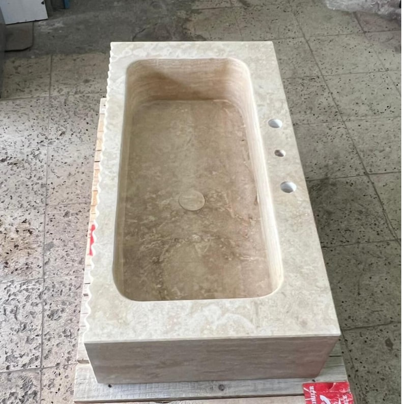 May include: A rectangular, beige stone bathtub with a rounded interior. The tub has a drain and three faucet holes. The exterior is a solid block of stone, and the tub is sitting on a wooden pallet.