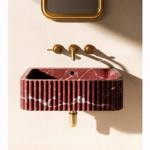 May include: A rectangular, red marble sink with vertical ribbed detailing. The sink has a white veining pattern and a brass drain. Above the sink are brass faucets and a gold-framed mirror.