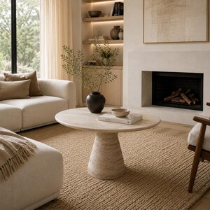 May include: A neutral-toned living room with a beige sofa, a round stone coffee table, and a woven rug. A fireplace and a wooden chair are also visible. Decorative items include a vase with branches and a bowl.