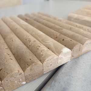 May include: A series of beige, semi-cylindrical stone tiles arranged in a parallel row. Each tile features a porous, textured surface with natural variations in color and light brown veining, creating a repeating, rounded linear pattern.