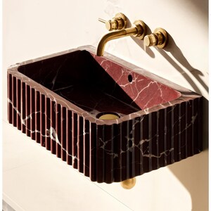 May include: A rectangular, wall-mounted sink made of red marble with white veining. The sink has a fluted exterior and a brass faucet and drain. The overall design is modern and luxurious, suitable for a bathroom.