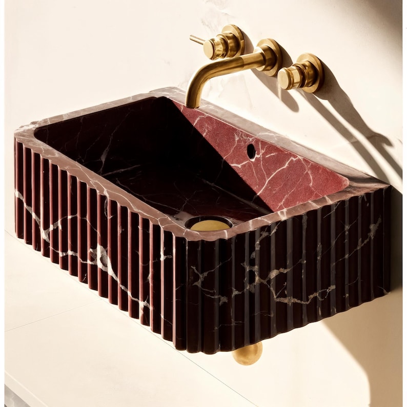 May include: A rectangular, wall-mounted sink made of red marble with white veining. The sink has a fluted exterior and a brass faucet and drain. The overall design is modern and luxurious, suitable for a bathroom.