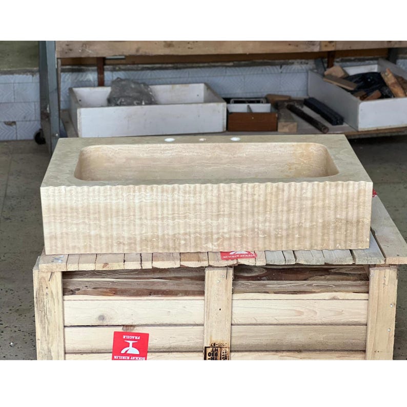 May include: A rectangular, beige stone sink with a textured, ribbed exterior. The sink has a smooth, recessed basin and three pre-drilled faucet holes. It rests on a wooden crate, with a red "Fragile" sticker visible.