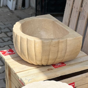 May include: A beige stone sink with a scalloped exterior. The sink has a rounded interior and is placed on a wooden crate. The sink's design features vertical grooves, adding texture to its surface. The overall aesthetic is rustic and natural.