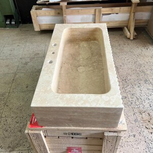 May include: A rectangular, beige stone sink with a textured surface. The sink has a basin, three faucet holes, and a decorative edge. It is sitting on a wooden crate.