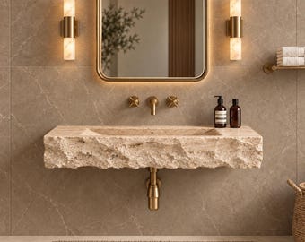 May include: A wall-mounted bathroom sink carved from beige natural stone with a rough, textured front edge. It features a brass faucet, two matching cross-handle valves, and a brass drain pipe. Two amber-toned glass bottles sit on the sink surface.