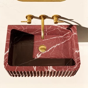 May include: A rectangular red marble sink with white veining. The sink has a gold faucet and fixtures. The front edge of the sink has a decorative, ridged design. The sink is a bathroom fixture.