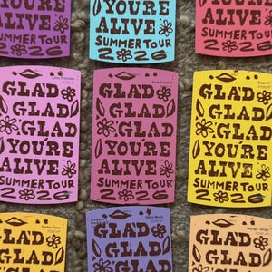 May include: A collection of rectangular stickers in various colors including purple, light blue, pink, yellow, and orange. Each sticker features the brown text GLAD YOU'RE ALIVE SUMMER TOUR 2026, decorated with small brown flower and leaf illustrations.