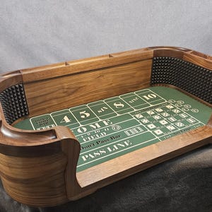 May include: A wooden craps table with a polished brown finish and a green felt surface featuring white printed betting layouts. The interior walls are lined with black pyramid-textured rubber bumpers for dice, and the top rail includes integrated chip slots.