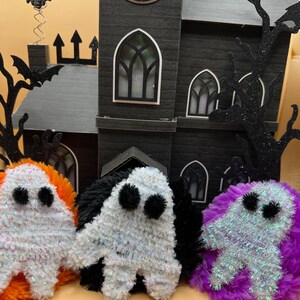 May include: A Halloween-themed display featuring three plush ghost figures in white, black, and iridescent fabric, with black pom-pom eyes. A black cardboard haunted house and tree silhouettes are in the background.