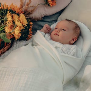 May include: A newborn baby wrapped in a soft, white swaddle blanket. The baby is wearing a white onesie and is nestled in a white, cushioned nest. A bouquet of yellow and orange flowers sits in a woven basket nearby.