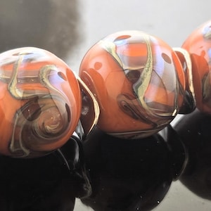May include: A strand of five orange and brown glass beads with swirling patterns. The beads are connected by silver-colored metal findings. The beads have a glossy finish and are reflected in the dark surface below.