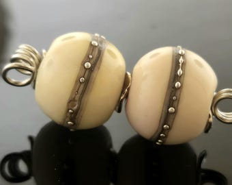 May include: Two white glass beads with silver accents and a silver wire connecting them. The beads have a striped pattern and are slightly different shades of white.