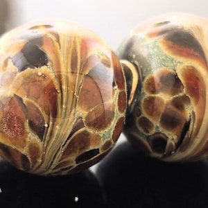 May include: Two artisan glass beads with swirling patterns in shades of brown, gold, and green. Each bead has a silver-colored metal loop attached. The beads are round and appear to be handmade, showcasing unique color variations and textures.