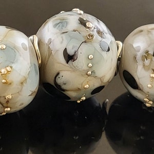 May include: A strand of six round glass beads with a mottled white, black, and gold pattern. The beads are strung on a silver wire.