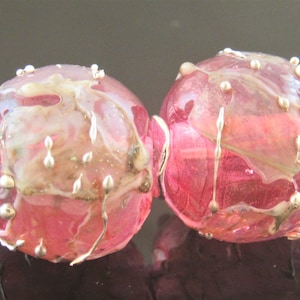 May include: Two pink glass beads with silver wire accents. The beads have a marbled effect with white and gold flecks.