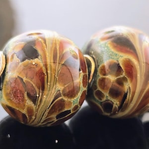 May include: Two artisan glass beads with a swirling pattern of brown, gold, and red hues. Each bead has a silver-colored metal loop attached. The beads are round and have a glossy finish.