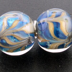 May include: Two glass beads with a swirling design in shades of blue, tan, and white. The beads are connected by a silver-colored metal finding. The glass beads have a glossy finish and are likely used for jewelry making.