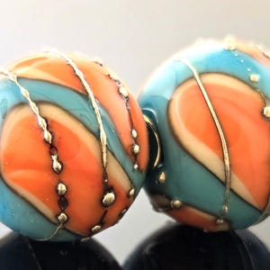 Puede incluir: Dos cuentas redondas de vidrio con un patrón de remolino de naranja, azul y blanco. Las cuentas están acentuadas con alambre dorado y pequeños puntos dorados.