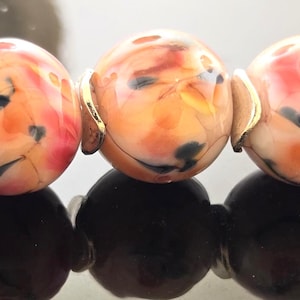 May include: A close-up of a bracelet featuring five speckled, round beads in shades of orange, red, and black. Each bead is connected with gold-colored wire. The bracelet's base consists of black beads.