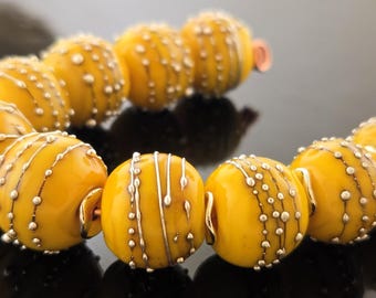 May include: A strand of yellow glass beads with silver and gold accents. The beads have a textured surface and are arranged in a circle.
