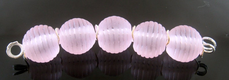 May include: A strand of six pink glass beads with a silver wire connecting them. The beads have a textured, ribbed surface.