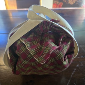 May include: A round, fabric lunch bag with a red and green plaid pattern. The bag is secured with a cream-colored strap that wraps around the top. The bag is sitting on a dark wooden surface.