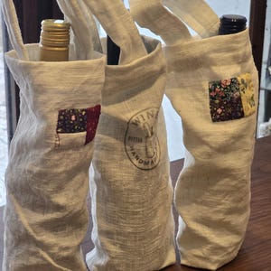 May include: Three off-white linen wine bottle bags with handles. Each bag has a small, colorful patchwork square on the front. One bag contains a gold-topped bottle, another a dark bottle, and the third is empty. The middle bag has a printed logo.