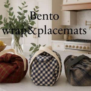 May include: Three bento boxes wrapped in fabric with handles. The boxes are in red plaid, blue and white gingham, and brown plaid patterns. The text "Bento wrap&placemats" is displayed above the boxes.