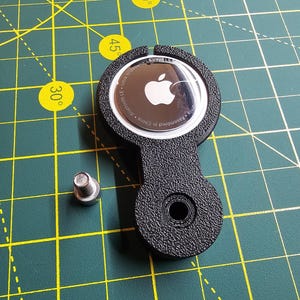 Puede incluir: Soporte negro para Apple AirTag con accesorio para llavero. El AirTag es visible con el logotipo de Apple. Un pequeño tornillo plateado está al lado del soporte. El fondo es una esterilla de corte verde con líneas de cuadrícula amarillas e indicaciones de grados.