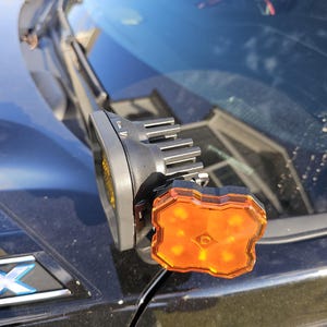 Puede incluir: Un accesorio de luz para vehículos negro y naranja montado en un vehículo negro. La luz tiene una lente ámbar rectangular con múltiples luces internas. El accesorio está sujeto a un soporte negro.