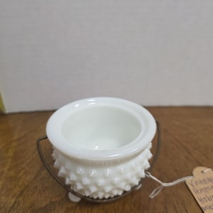 May include: A white hobnail milk glass bowl with a metal handle. The bowl has a round opening and a textured exterior. A small tag is attached to the handle.