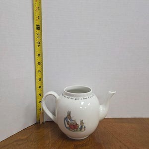 May include: White ceramic teapot with a round body, a curved handle, and a spout. The teapot features a rabbit illustration and text. Dimensions: 6" wide, 5.5" high, and 9" deep.