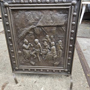 May include: A rectangular bronze fireplace screen featuring a raised relief scene of people gathered outside a cottage. The frame is decorated with a border of circular studs and includes a central handle at the top and two metal feet at the base.