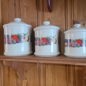 Puede incluir: Juego de tres botes de cerámica blanquecina con tapas. Cada bote presenta una colorida banda de ilustraciones de frutas, incluyendo uvas, manzanas y peras. Los botes se exhiben en una estantería de madera.